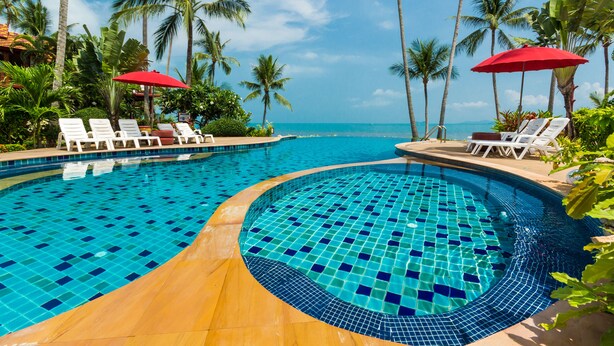 A pool with tiled edges, surrounded by palm trees and lounge chairs with red umbrellas.