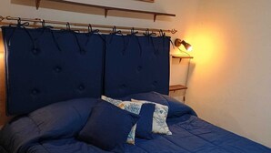 A bedroom with a large bed, a headboard, a wall-mounted shelf, a painting, and a bedside lamp. - Apartment/ flat - Levanzo (Levanzo)