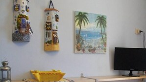 A small kitchen area with two wooden chairs, a yellow cloth on one, a black monitor on a shelf, and a painting of a beach scene on the wall. - Apartment/ flat - Levanzo (Levanzo)