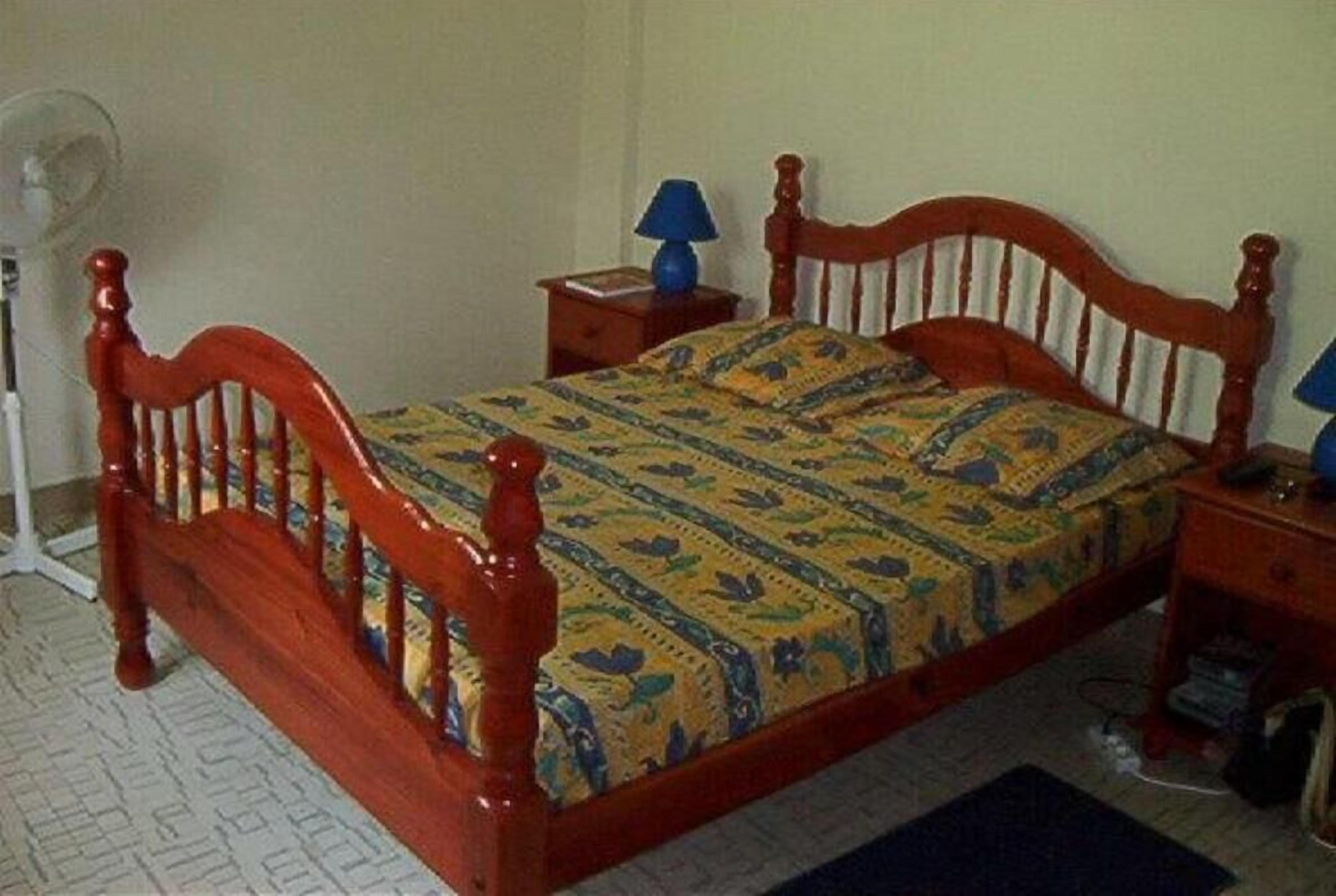 A single bed with a wooden frame and a patterned bedspread. There is a small bedside table with a lamp and a book. A floor lamp is visible in the corner.