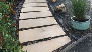 Een pad dat leidt naar een gebouw met een groen en wit gevel, een bankje en bloempotten.
