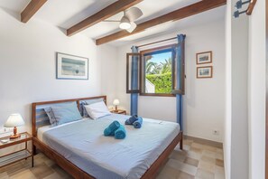 Ein Schlafzimmer mit einem Bett, einem Nachttisch, einer Lampe, einem Deckenventilator und einem Fenster mit Blick auf Grünflächen.