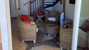 A cozy living area with wicker furniture, a red cushion, a wooden chair, and a small table with books and a fan.