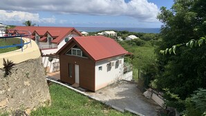 A small house with a red roof and white walls, surrounded by lush greenery and a view of the ocean.