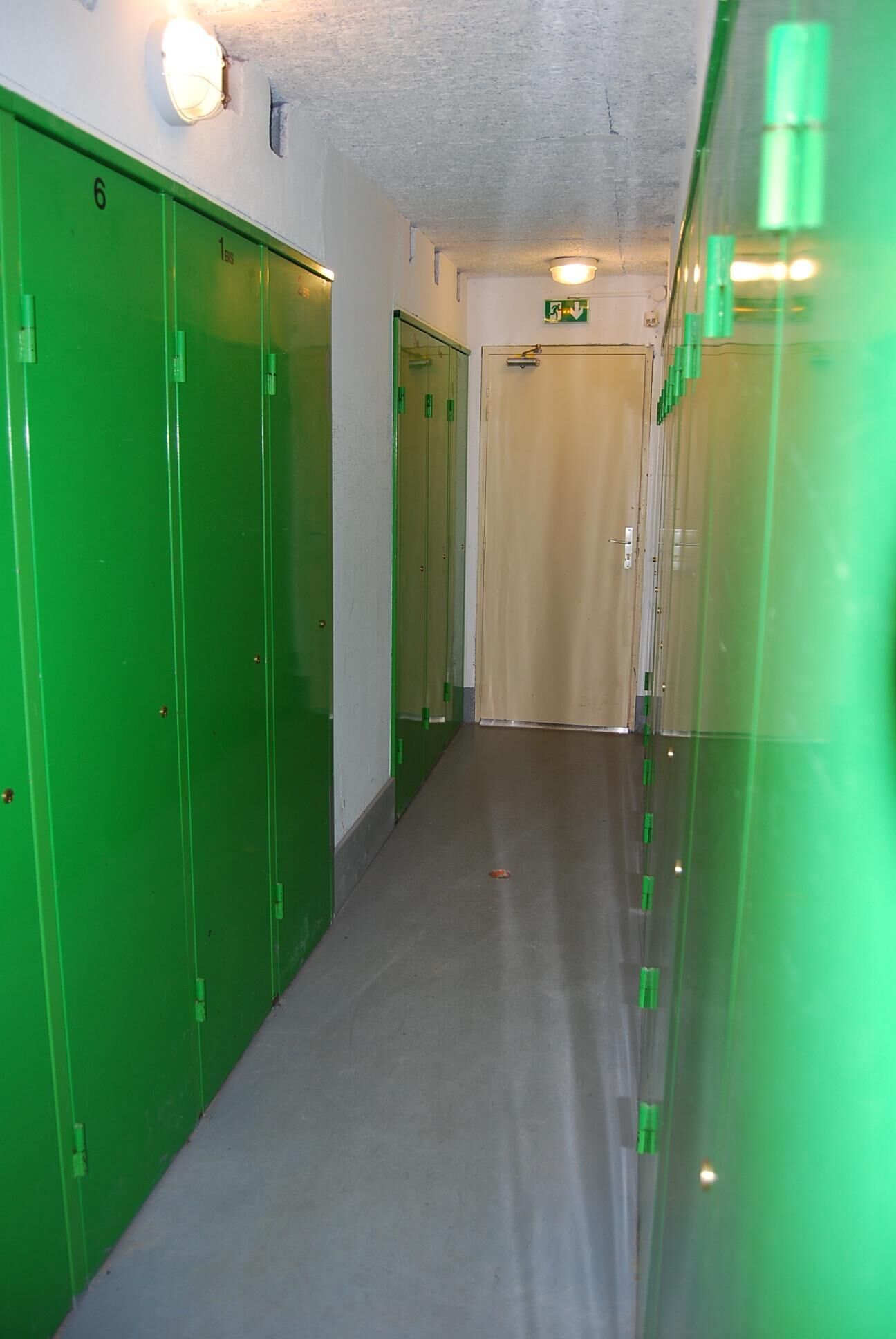 Un pasillo con taquillas verdes, una puerta color crema y un letrero de salida de emergencia verde.