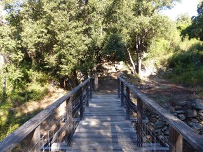 Un pont en bois doté d’une rambarde métallique, entouré d’arbres denses et bordé d’un mur de pierre d’un côté.