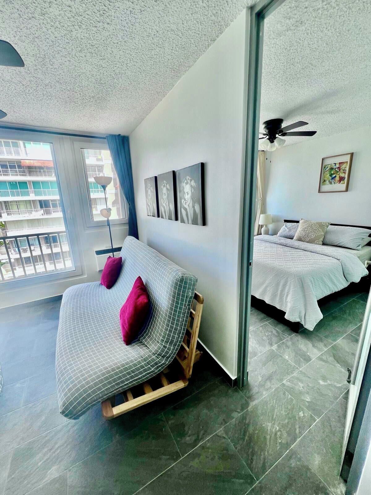 A modern hotel room with a large bed, a smaller sofa, and a balcony with a view of the city.