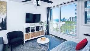 A modern living room with a flat-screen TV, a ceiling fan, a large window with a city view, a sofa, and a small round coffee table. - Flash Sale!!!  Newly Renovated Oceanview Property in Isla Verde #2! (Carolina)