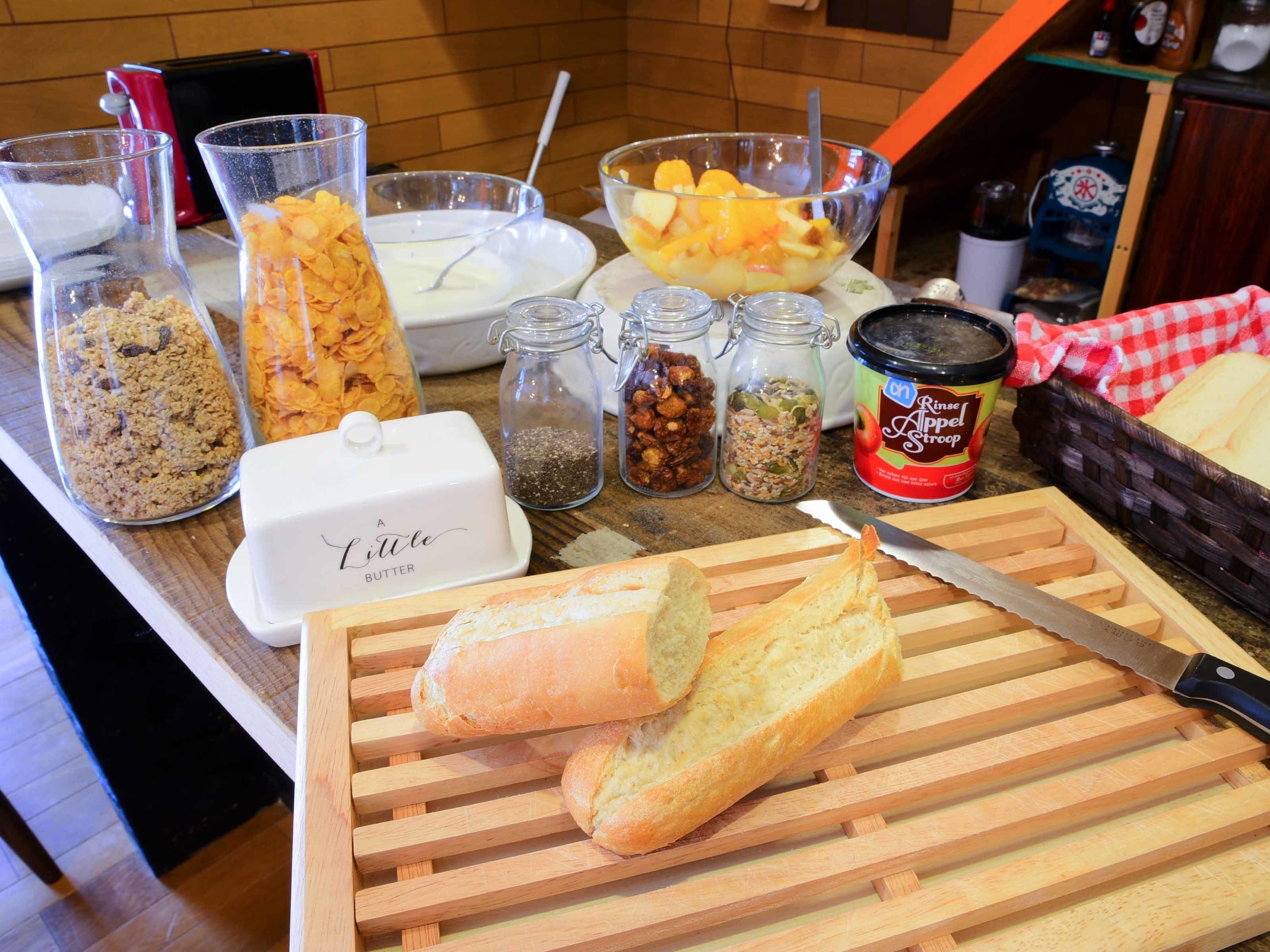 A kitchen counter with various food items and utensils, including jars of spices, a bowl of fruit, and a loaf of bread.