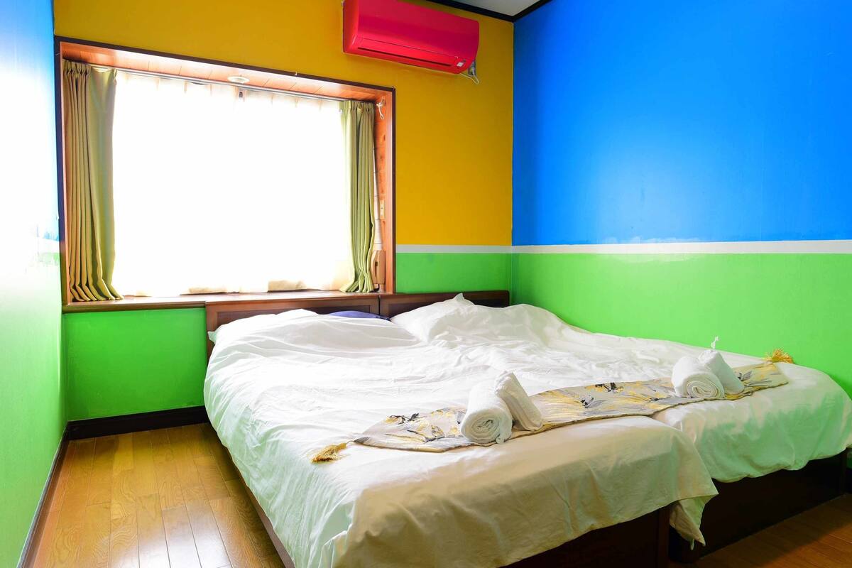 Una habitación con una cama, un ventanal con cortinas, un aire acondicionado rojo y una pared pintada en tonos amarillo, verde y azul.