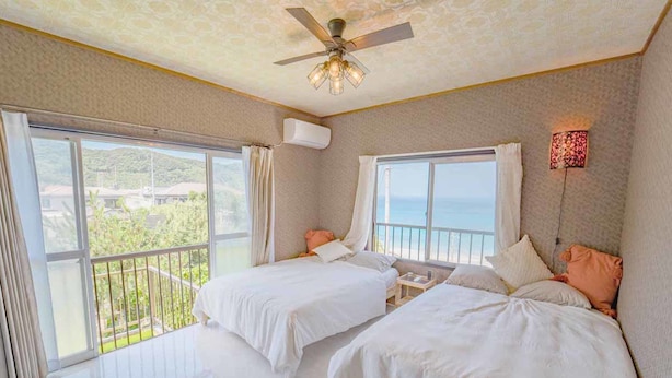 Habitación de hotel con dos camas, ventilador de techo y ventanas amplias con vista al mar.