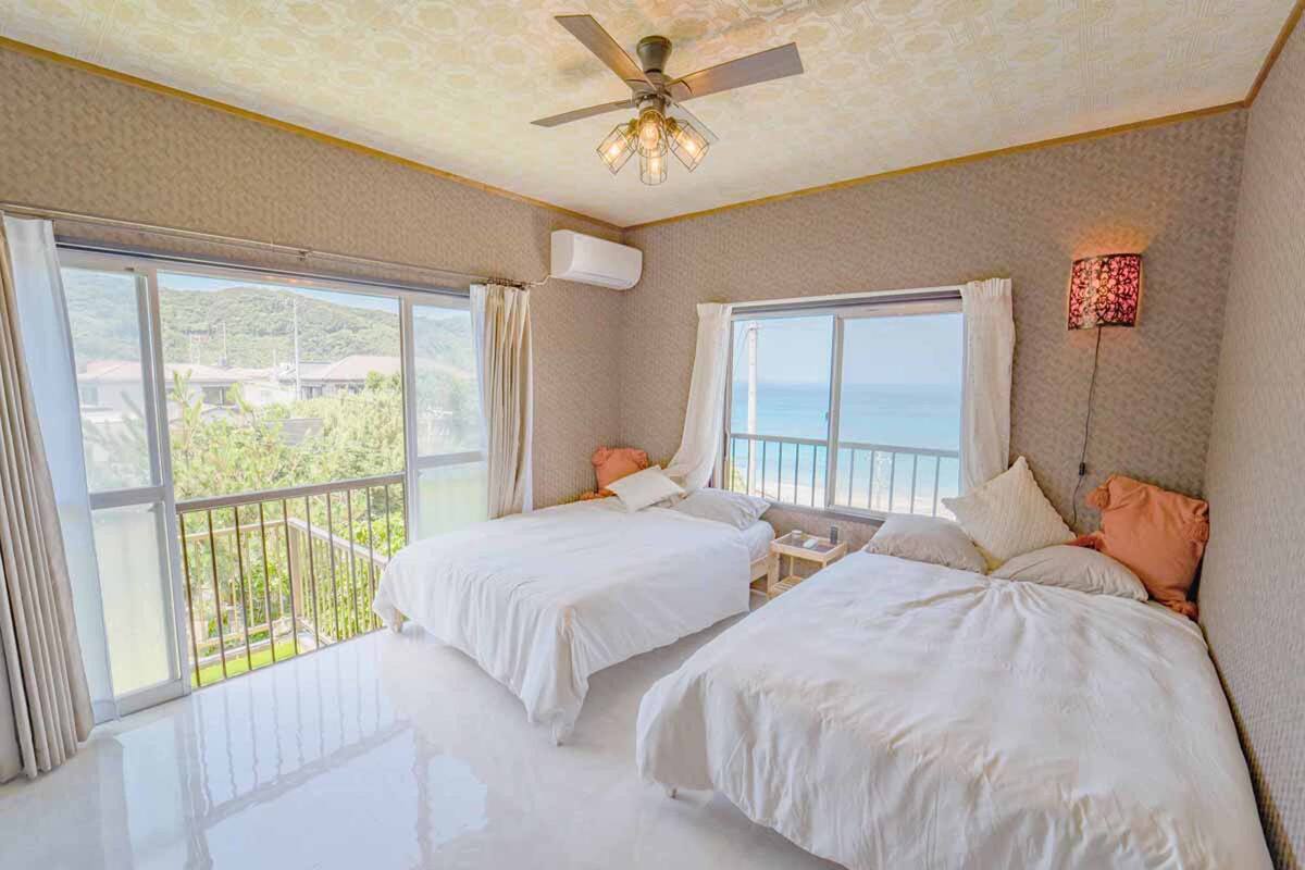 Habitación de hotel con dos camas, ventilador de techo y ventanas amplias con vista al mar.