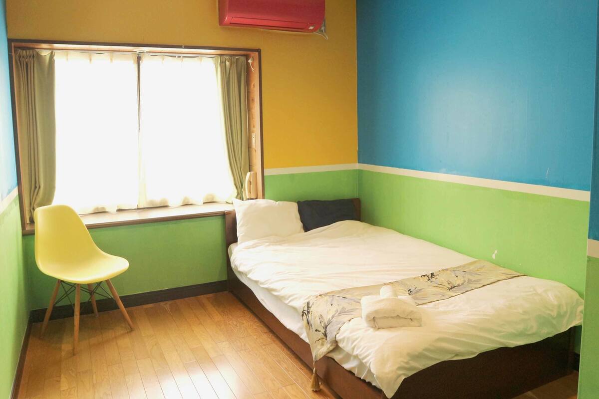 Una habitación pequeña con cama individual, ventana, una silla y aire acondicionado instalado en la pared.