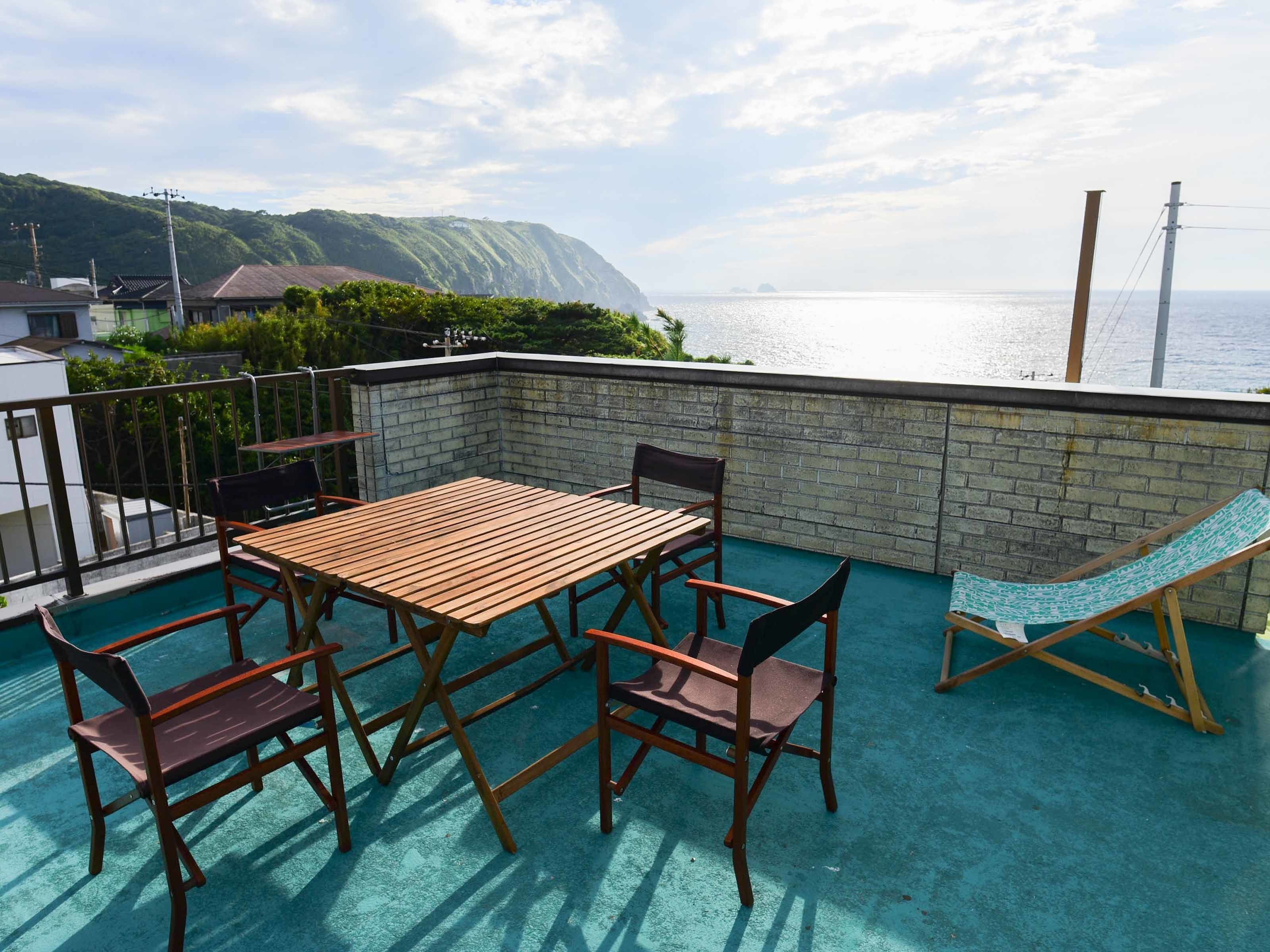 A rooftop terrace with a wooden table and chairs, a hammock, and a view of the sea and hills.