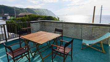 A rooftop terrace with a wooden table and chairs, a hammock, and a view of the sea and hills.