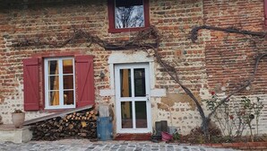 Um edifício rústico de tijolo com telhado de telhas, uma pequena janela com vista para árvores sem folhas, uma porta com moldura branca e um pátio pavimentado em pedra.