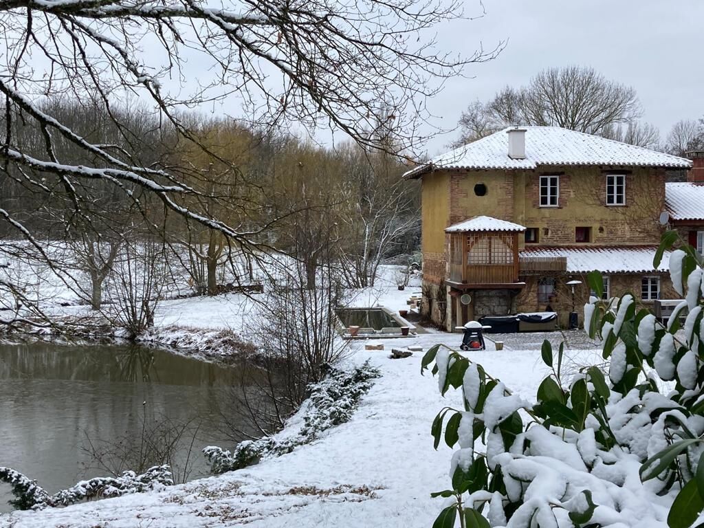 Une maison de campagne à l’aspect rustique, avec une façade en pierre et un toit recouvert de neige, située au bord d’une rivière.