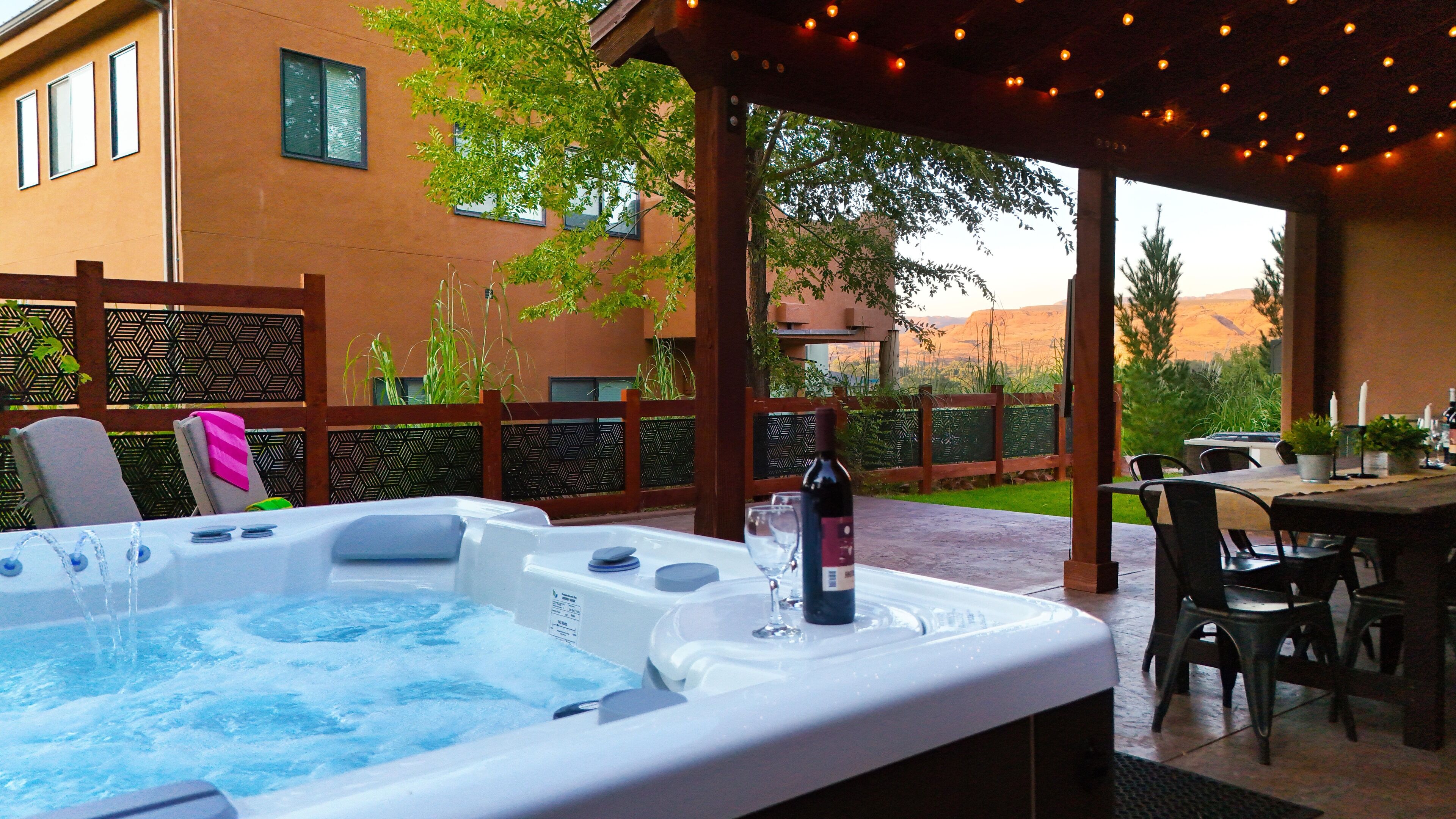 A hot tub area with a table and chairs, surrounded by a wooden pergola and string lights.