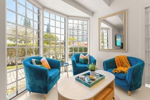 Ein modernes Wohnzimmer mit großen Fenstern, zwei blauen Sesseln, einem Couchtisch mit Dekoration und einem Spiegel an der Wand.