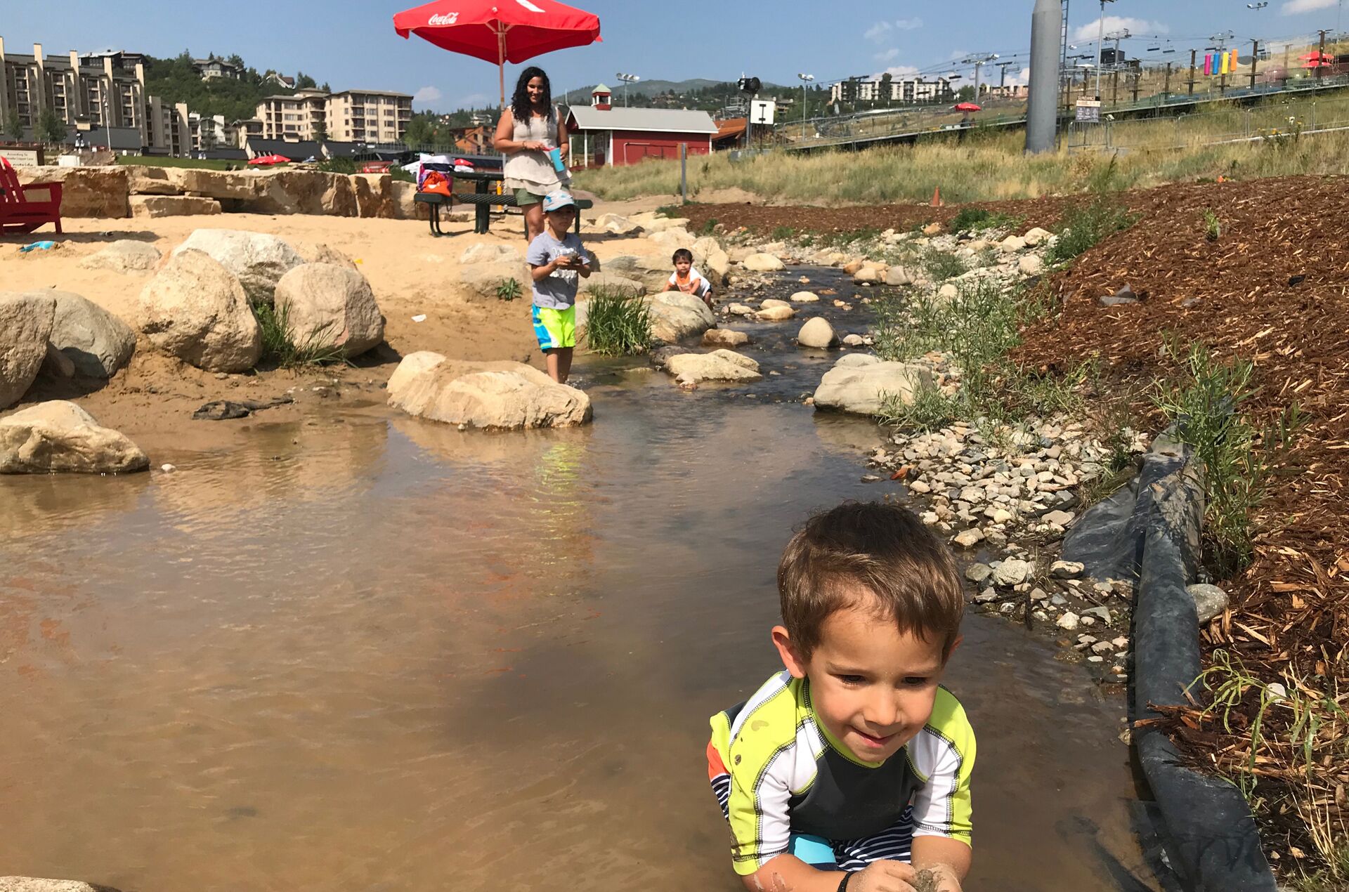 A child playing in a shallow stream with rocks and a woman in the background.
