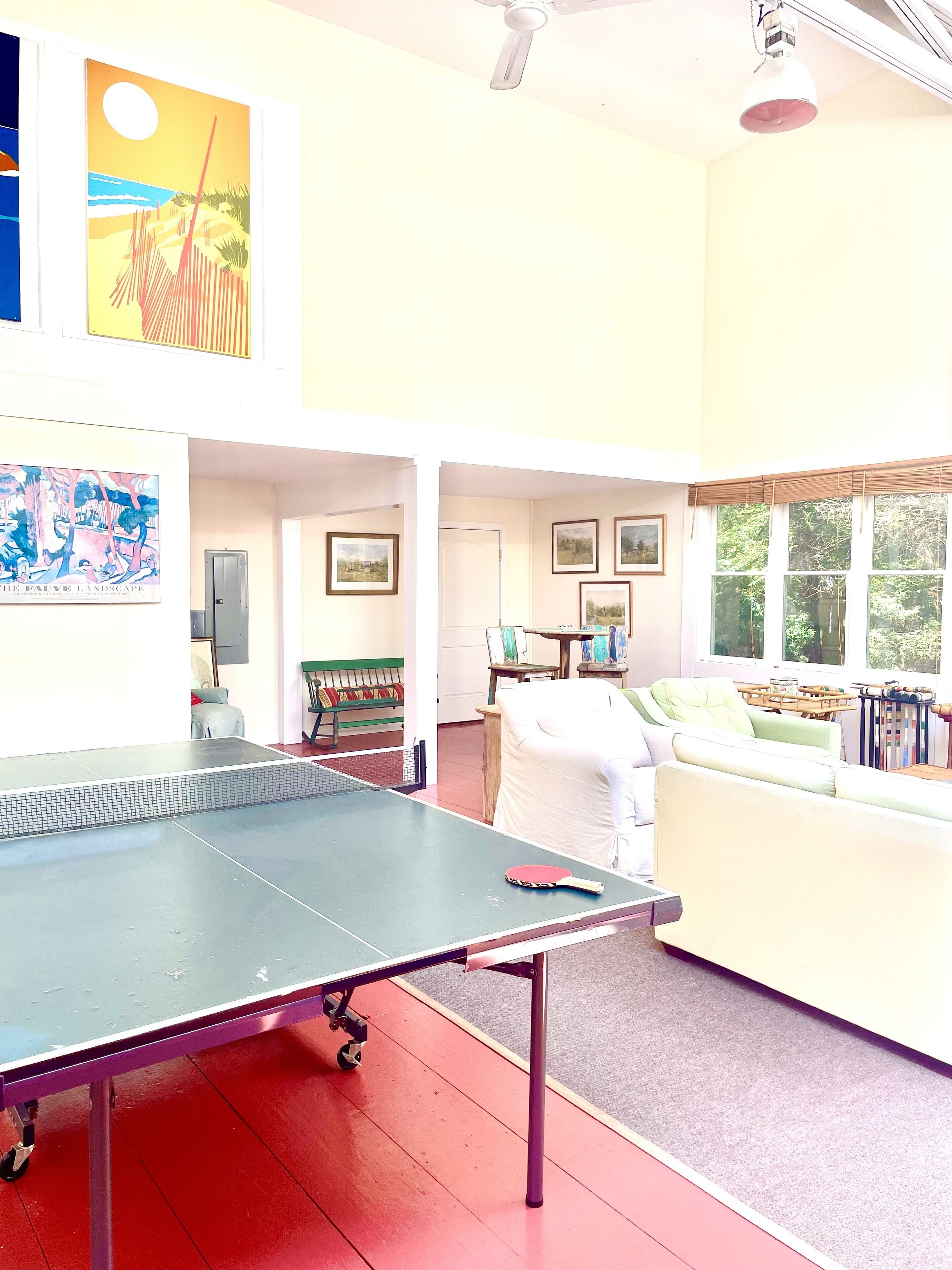 A room with a ping pong table, a sofa, and a window with a view of trees.