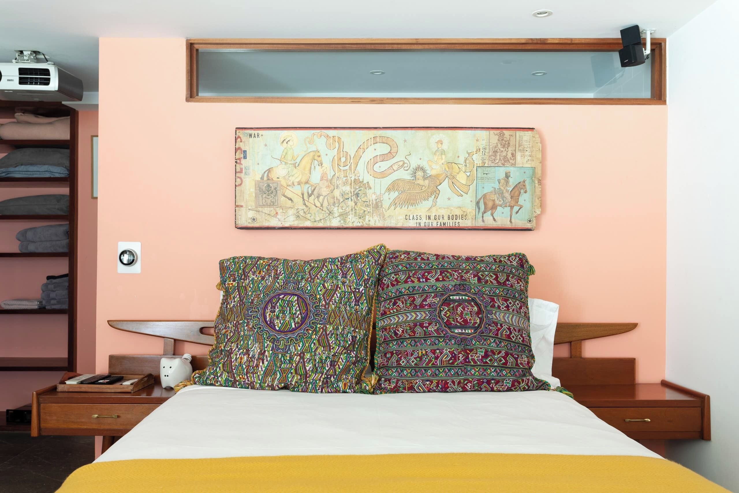 Un dormitorio con cama, mesita de noche, lámpara, estante con mantas dobladas y proyector instalado en la pared.