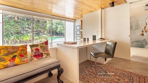 Een moderne kamer met een houten plafond, een groot raam met uitzicht op groen, een wit bureau, een zwarte stoel en een kamerbreed tapijt met een patroon.