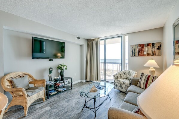 A modern living room with a flat-screen TV, wicker furniture, a glass coffee table, and a large window offering a view. - Modern and Clean Condo, Indoor and Outdoor Pools | Waterpointe II - 302 (North Myrtle Beach)