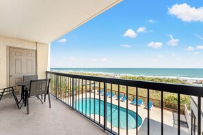 En balkon med udsigt over strand og hav, en pool samt udendørs siddepladser.