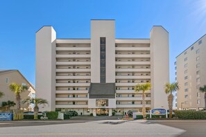 A multi-story hotel with a central entrance, parking area, and palm trees.