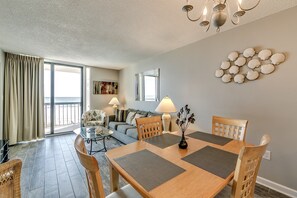 A dining area with a wooden table, chairs, and a chandelier. There is a sofa and a coffee table in the background. - Modern and Clean Condo, Indoor and Outdoor Pools | Waterpointe II - 302 (North Myrtle Beach)