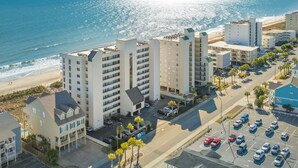 A coastal hotel with multiple buildings, a parking lot, and a beach view.