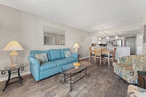 Un salon avec un canapé bleu, une table basse et un fauteuil à motifs. On y trouve également un coin repas avec une table et des chaises en bois. La cuisine est visible au fond, avec des meubles blancs et des appareils en acier inoxydable.