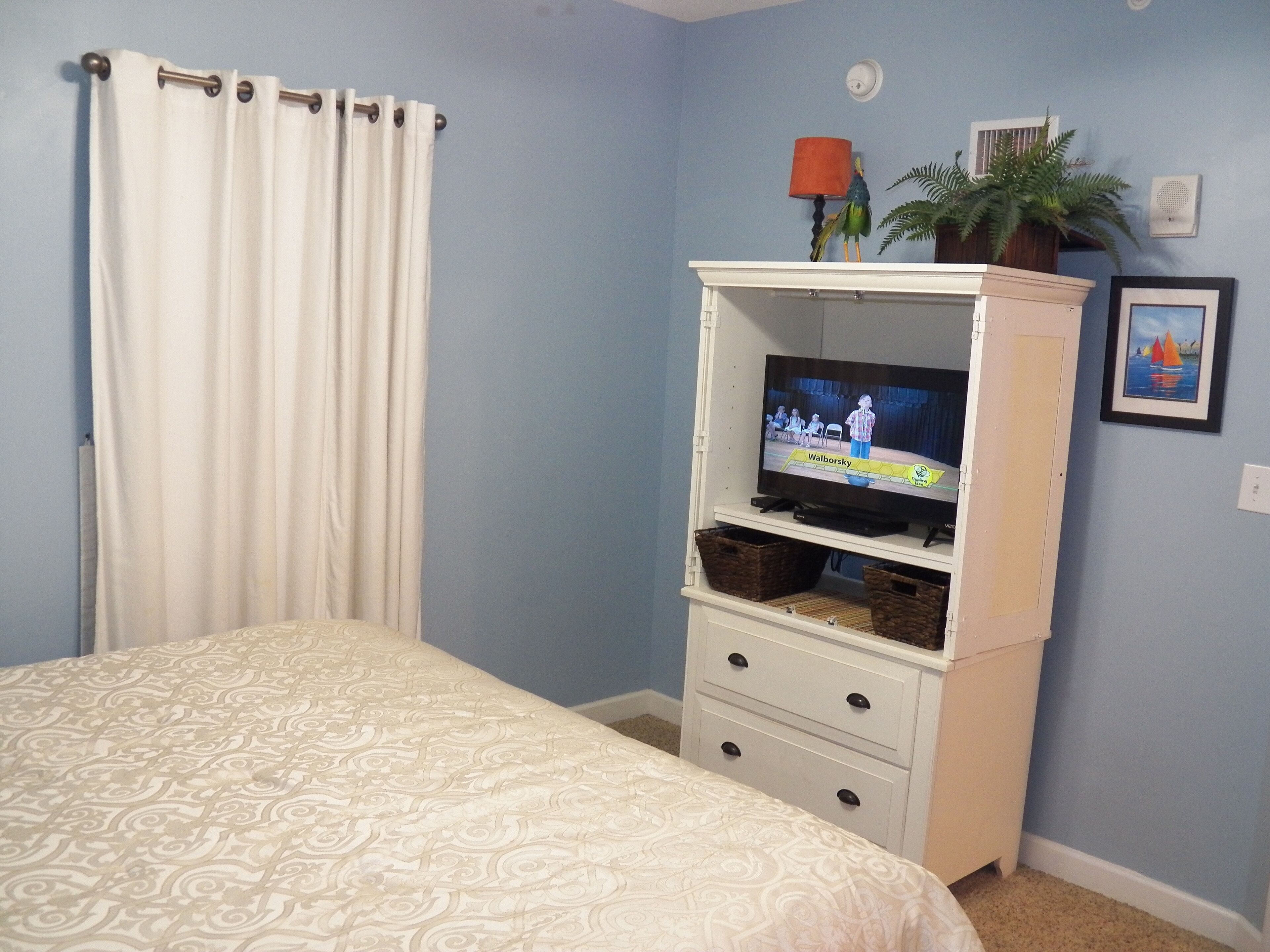 Quarto com cama, televisão sobre um suporte, cómoda branca e janela com cortinas.