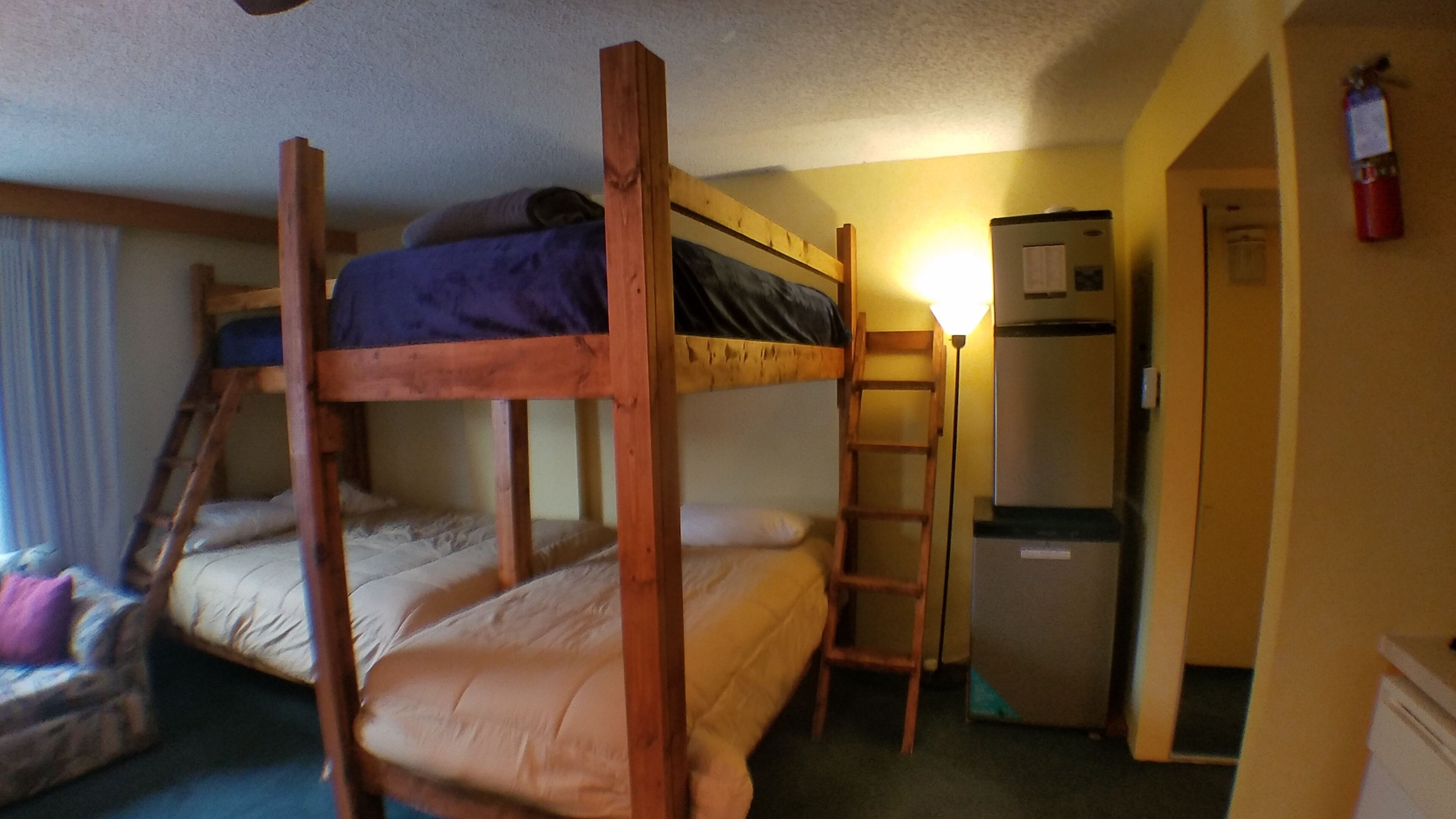 A bunk bed room with a ladder, a refrigerator, and a door leading to another room.