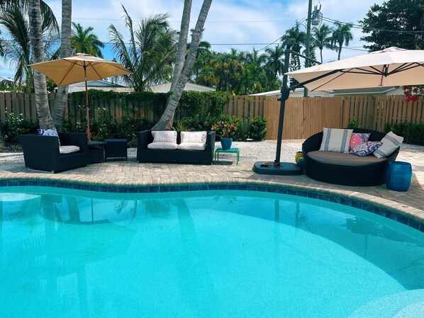 A pool area with lounge chairs and umbrellas, surrounded by palm trees and a wooden fence.