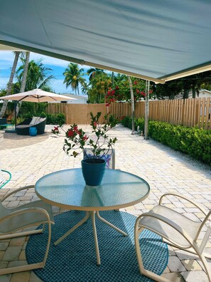 An outdoor patio with a round glass table, two chairs, a blue potted plant, and a wooden fence in the background. - Pet Friendly Beachy Home in the Heart of Naples! Multi Month Discount! (Naples)