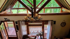 L'intérieur d'une cabane avec un plafond en bois, un canapé en cuir, un lustre et une vue sur l'extérieur grâce à de grandes fenêtres.