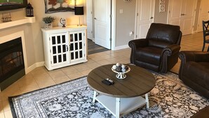 Una sala de estar con chimenea, un televisor montado encima, un mueble blanco con puertas de cristal, un sillón de cuero negro y una alfombra estampada.