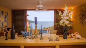 A room with a view of the ocean, a ceiling fan, a table with candles and decorative items, and a vase with white flowers. - The Beach is Calling!  Book November & December NOW! (Zihuatanejo)