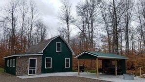 A green building with a stone foundation, a covered outdoor area, and a gravel driveway. - Petoskey - Enjoy the great outdoors from the Cozy and peaceful cabin! (Petoskey)