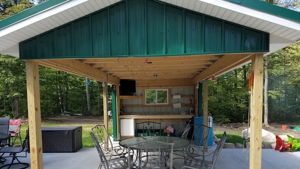 A covered outdoor patio with a table and chairs, surrounded by greenery. - Petoskey - Enjoy the great outdoors from the Cozy and peaceful cabin! (Petoskey)