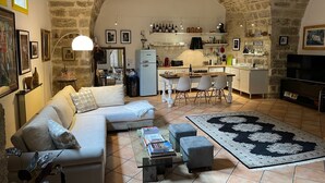 Un salon chaleureux avec un canapé, une table basse et des poufs, une cuisine équipée d’un réfrigérateur et d’un coin repas, et un plafond en pierre.