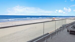 A beach view from a balcony with a railing, a lifeguard chair, and a small seating area.
