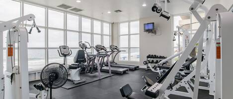 Un gimnasio con equipo de ejercicio diverso, ventanas amplias y un ventilador.