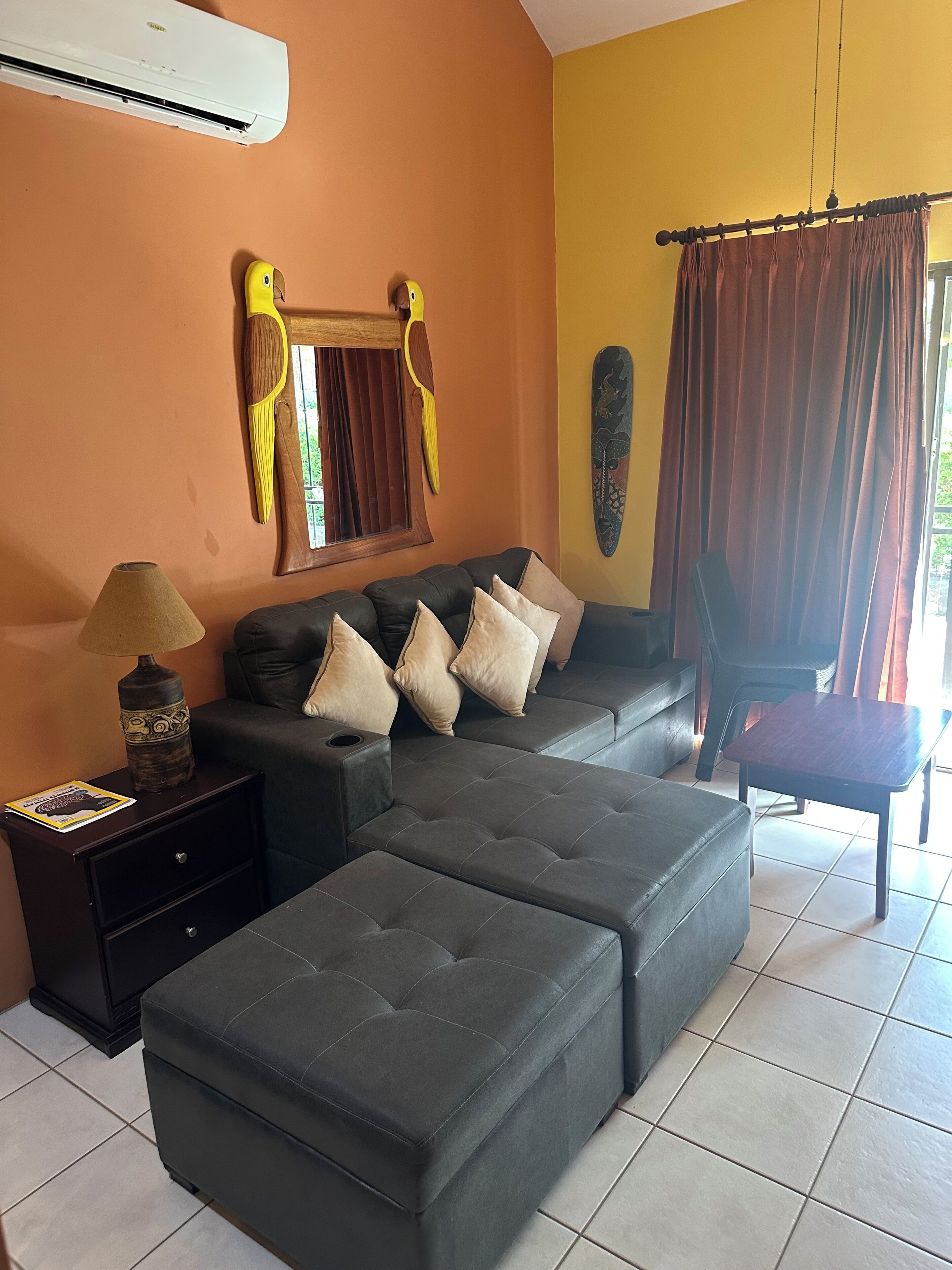 A living room with a sectional sofa, a small table, a lamp, and a wall-mounted air conditioner.