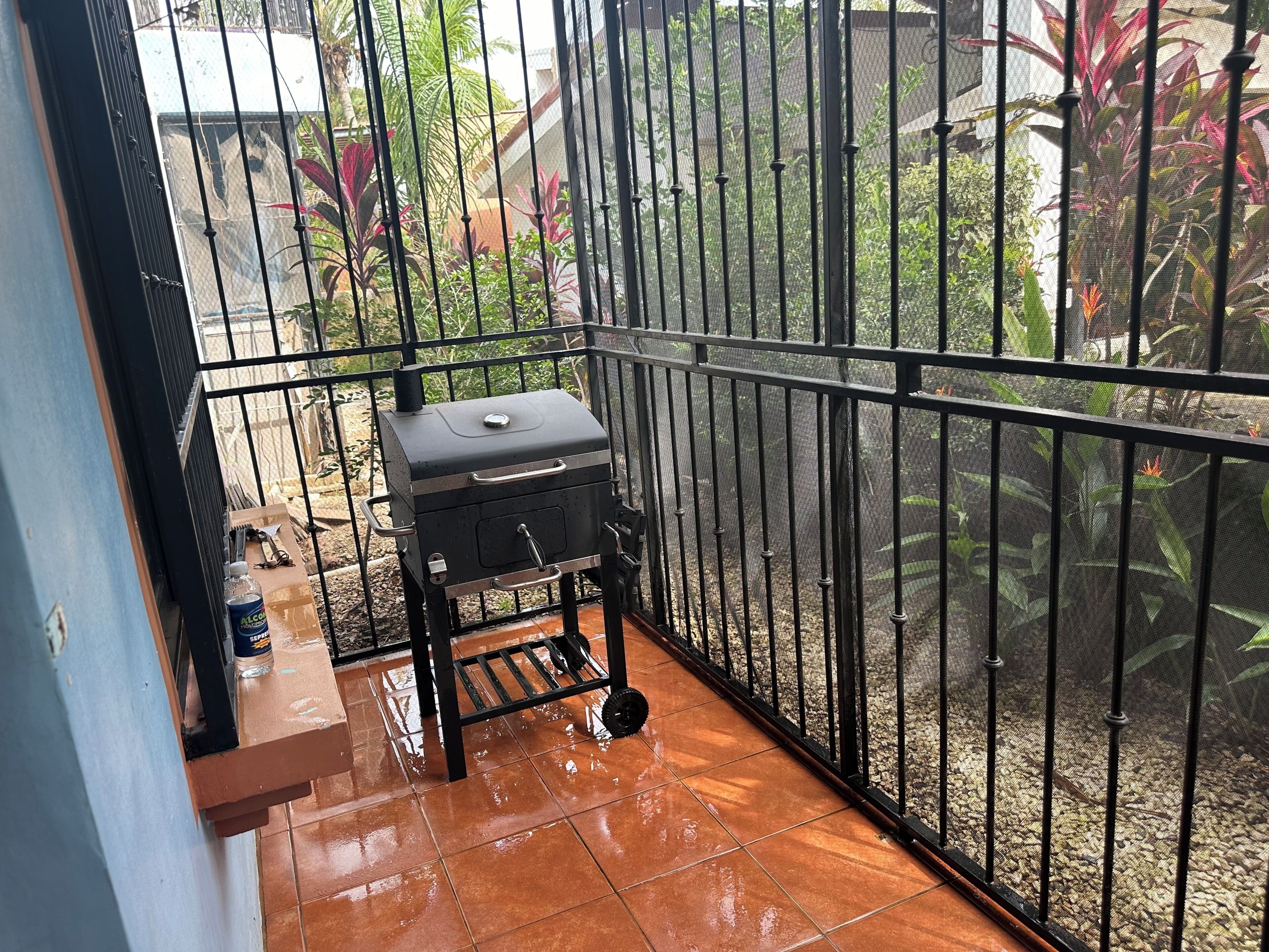 Un balcón con una parrilla, una mesita con una botella y una silla.