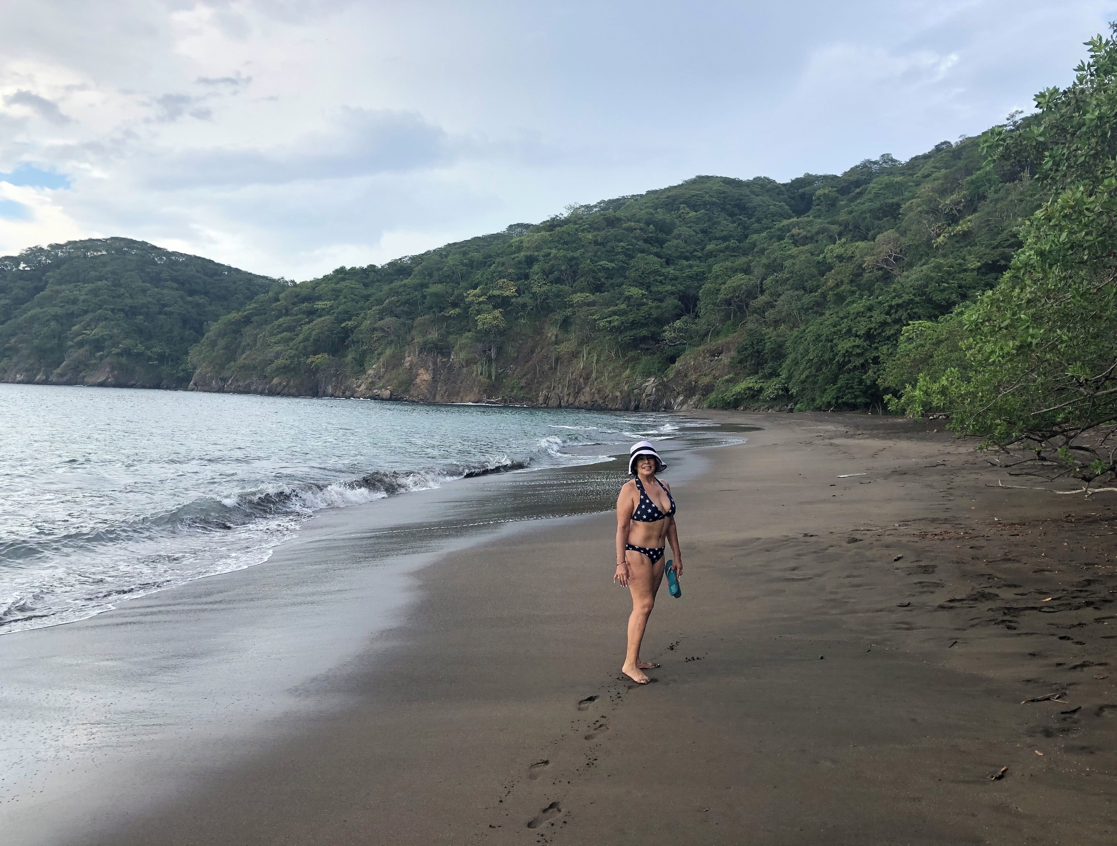 Una playa con una persona en traje de baño y sombrero, rodeada de vegetación y colinas.