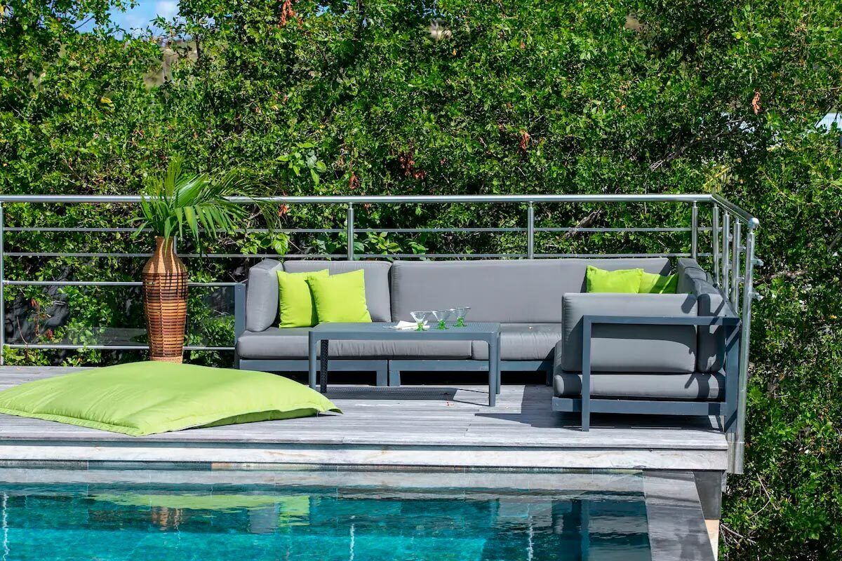 Un espace aménagé au bord de la piscine, avec un canapé gris, des coussins verts, une plante en osier et une rambarde en métal.