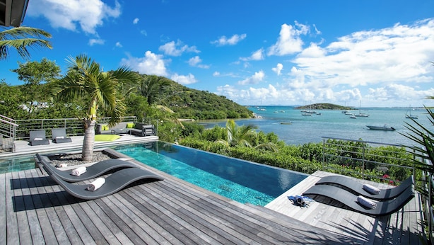 A poolside area with lounge chairs, a palm tree, and a view of the sea with boats. - Luxury Waterfront Villa Pinel. Direct Ocean Views & Private Beach Access (SAINT MARTIN)